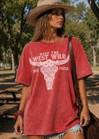 A woman wearing a faded red graphic t-shirt with a white longhorn bull design and the text 'KEEP THE WEST WILD', paired with a cowboy hat and denim shorts, standing outdoors in a park setting.
