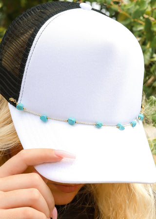 White trucker cap with black mesh back and turquoise beads held by a person outdoors.