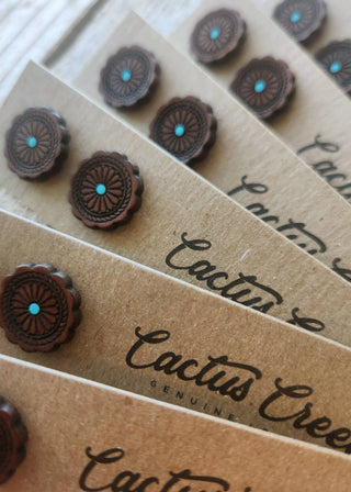 Close-up of several brown kraft paper cards printed with the brand name 'Cactus Creek Genuine' and each topped with a matching dark brown flower-shaped stud earring with a small turquoise center, arranged in a fan on a light wood surface.