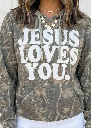 A woman wearing a camouflage hoodie with the words 'JESUS LOVES YOU.' printed in large white letters across the front, paired with white pants.