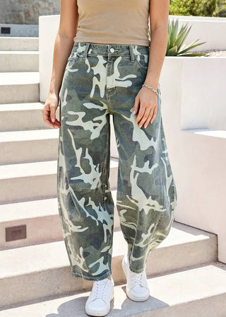 A person wearing loose-fit camouflage cargo-style jeans with a green, gray, and off-white camo pattern, paired with a beige tank top and white sneakers, standing on outdoor concrete steps.