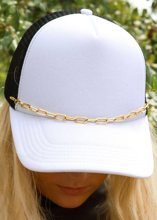 White cap with a gold chain worn by a person against a green leafy background