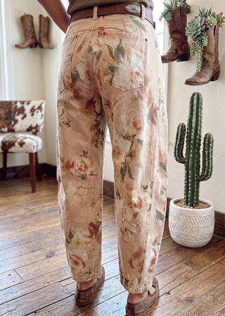 A person shown from the back wearing floral wide-leg jeans with a brown belt and brown shoes, standing on a wooden floor in a room with cowboy boots and a cactus.