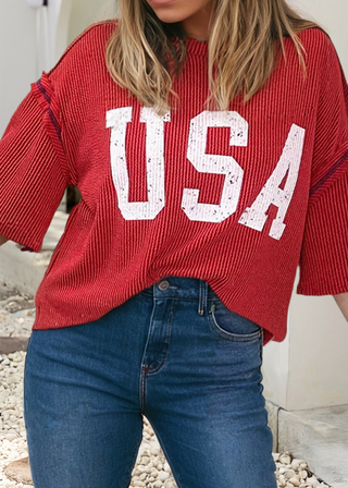 Woman wearing a red ribbed top with bold white “USA” lettering and red stitching