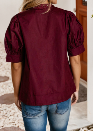 A woman viewed from the back wearing a burgundy short-sleeve blouse with puff sleeves and blue jeans, standing in a doorway with a pebble ground.